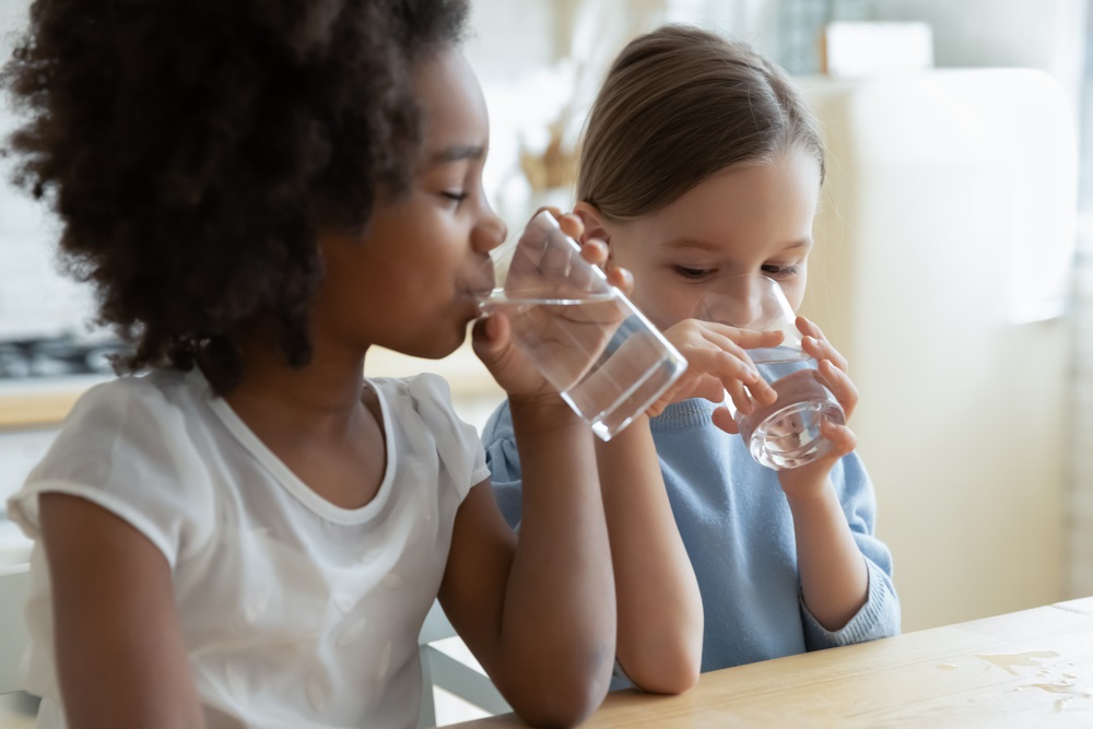 Two young girls drink water out of clear glasses at a kitchen table
