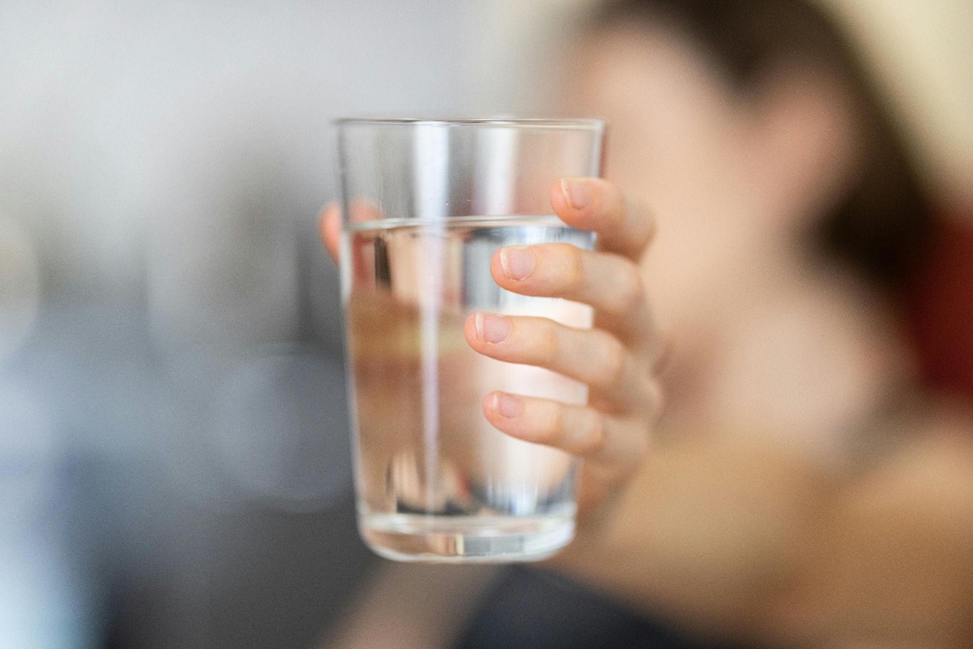 A woman's hand holds a glass of clean drinking water to the camera, with the woman out of focus in the distance