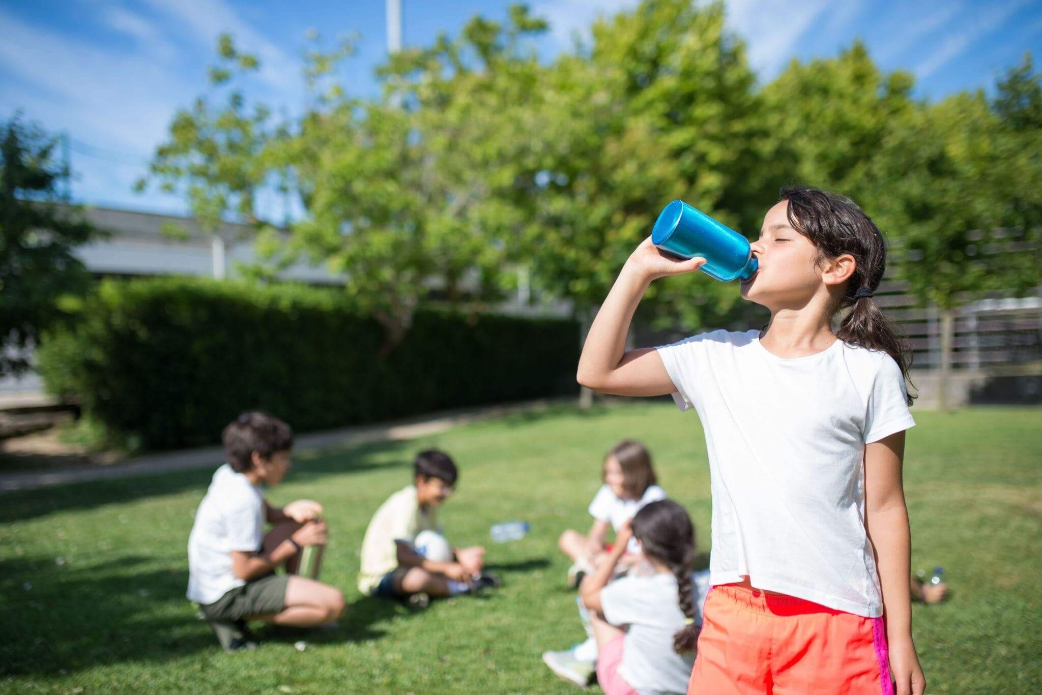 Group of children playing at an outdoor event with a young girl in the foreground drinking from a reusable water bottle