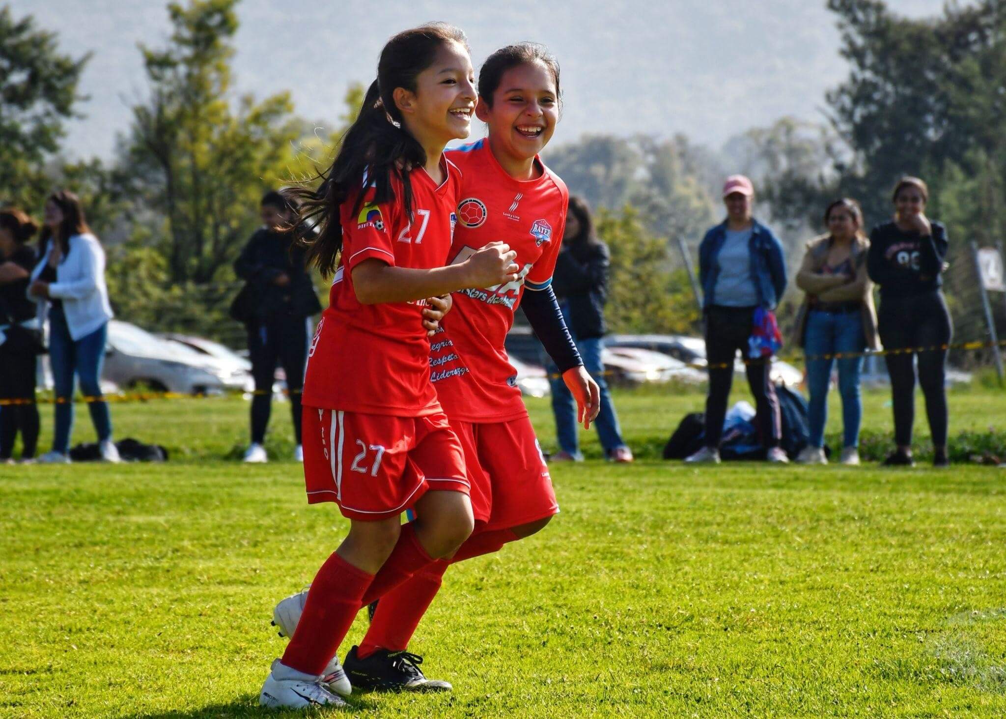 Two young girls playing soccer at a tournament as a group of parents and family watches from the sidelines